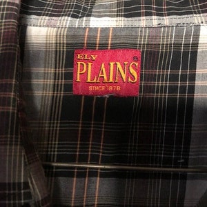 May include: Close-up of a plaid button-down shirt. The shirt features a pattern of black, grey, white, and orange lines. A red tag with the text "ELY PLAINS SINCE 1878" is sewn on the shirt.