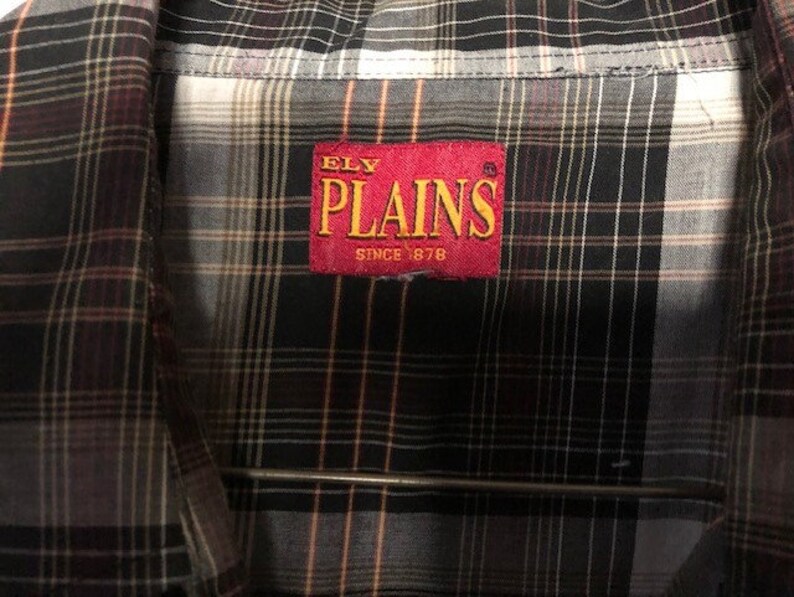 May include: Close-up of a plaid button-down shirt. The shirt features a pattern of black, grey, white, and orange lines. A red tag with the text "ELY PLAINS SINCE 1878" is sewn on the shirt.