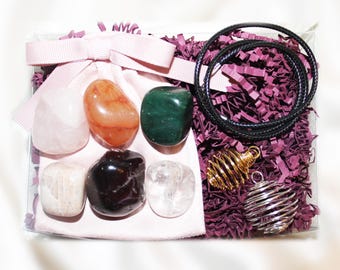 May include: A clear gift box containing a light pink drawstring pouch with six polished tumbled stones in shades of white, orange, green, black, and clear. Includes two metal spiral pendant cages and a black cord necklace on purple paper filler.