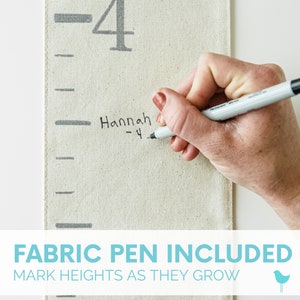 May include: A white fabric growth chart with black markings and the number 4 printed on it. A hand is writing "Hannah - 4" on the chart with a black fabric pen. The text "FABRIC PEN INCLUDED MARK HEIGHTS AS THEY GROW" is printed below the chart.