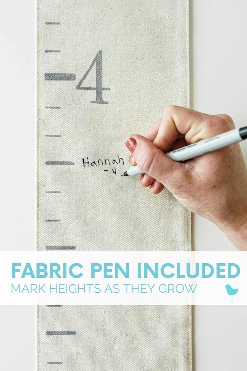 May include: A white fabric growth chart with black markings and the number 4 printed on it. A hand is writing "Hannah - 4" on the chart with a black fabric pen. The text "FABRIC PEN INCLUDED MARK HEIGHTS AS THEY GROW" is printed below the chart.
