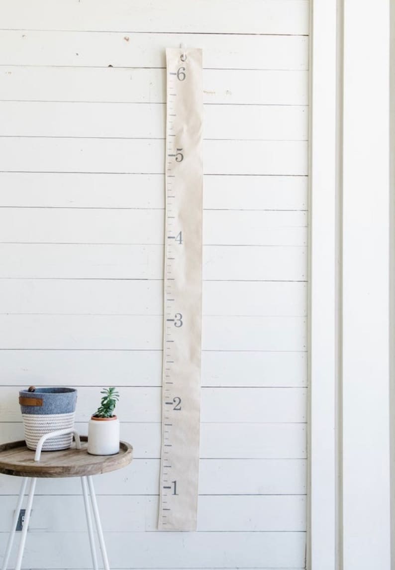 May include: A beige fabric growth chart with black numbers from 1 to 6 hanging on a white wood wall. The chart is a simple design with a straight line and numbers on the left side.