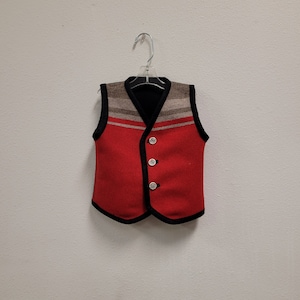 May include: A red vest with a black trim and three decorative buttons. The upper portion features a striped pattern in shades of brown and gray. The vest is hanging on a clear plastic hanger against a white wall.