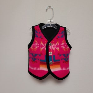 May include: A pink fleece vest with a black trim and a geometric pattern in shades of blue, orange, and white. The vest has three silver buttons.