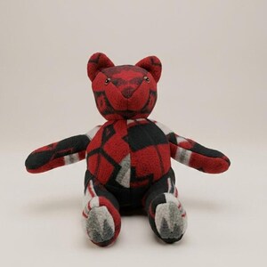May include: A plush teddy bear constructed from fleece fabric featuring a geometric pattern in shades of red, black, and grey. The bear has small dark eyes, rounded ears, and a seated posture with outstretched arms, showcasing a patchwork design.