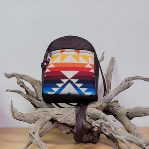 May include: A dark brown leather backpack with a colorful geometric design on the front pocket. The design features a pattern of triangles and stripes in shades of orange, red, blue, and white. The backpack has a top handle and adjustable straps.