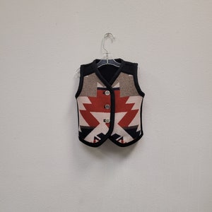 May include: A black vest with a geometric pattern in red, white, and blue. The vest has a button closure and is hanging on a hanger.