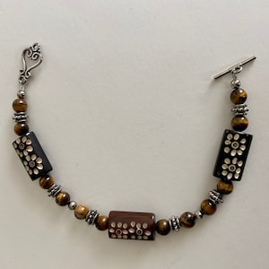 May include: A brown and black beaded bracelet with silver accents. The bracelet features three rectangular beads with white floral designs. The bracelet has a silver toggle clasp.