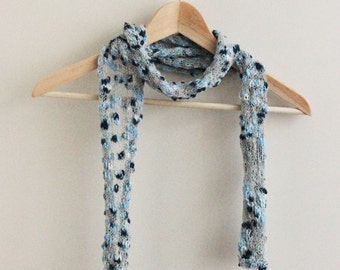 Peut inclure: Une écharpe en tricot bleu clair et gris avec un motif texturé de petits points bleus et blancs. L'écharpe est suspendue à un cintre en bois.