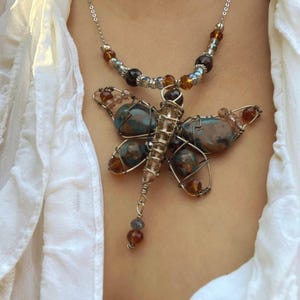May include: A handcrafted necklace featuring a dragonfly pendant. The dragonfly has blue and brown stone wings, a wire-wrapped body, and a beaded chain. The necklace is worn over a white shirt.