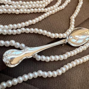 May include: A silver spoon rests amidst multiple strands of white pearl necklaces. The spoon has an ornate handle design. The pearls are small and round, creating a classic jewelry aesthetic. The background is a solid brown fabric.