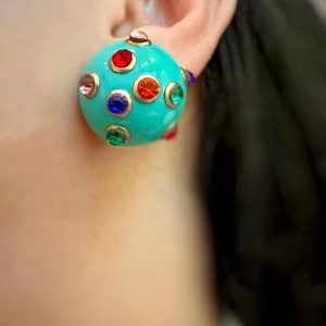 以下が含まれることがあります： カラフルな宝石のようなアクセントが付いた、ターコイズ色の丸いイヤリング。イヤリングには、それぞれ異なる色の宝石を保持する、一連の小さな丸い金色のセッティングがあります。宝石には、赤、オレンジ、ピンク、青、緑が含まれます。