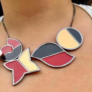 Könnte beinhalten: Eine geometrische Halskette mit einer silbernen Kette und Anhänger. Der Anhänger zeigt Formen in Rot, Schwarz und Gelb. Die Halskette wird über einem weißen Hemd getragen.