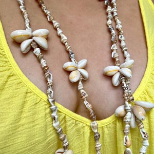 May include: A multi-strand necklace featuring natural seashells in shades of white, tan, and brown. The necklace has a layered design with larger shell accents and is worn against a yellow top. The jewelry is a beach-inspired accessory.