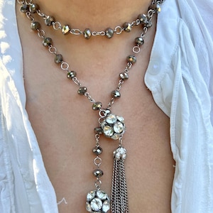 Puede incluir: Un collar de varias capas con dos hilos de cuentas grises facetadas. El collar presenta dos colgantes grandes y redondos con incrustaciones de cristal y borlas de cadena plateada colgantes. El collar se lleva sobre una blusa blanca.