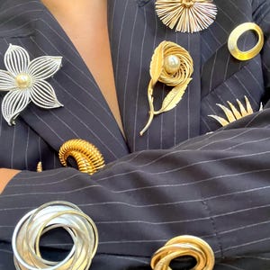 May include: A collection of vintage brooches adorns a navy blue pinstripe blazer. The brooches include a silver flower with a pearl center, gold floral and geometric designs, and a gold spiral brooch. The blazer's fabric is dark with thin white stripes.