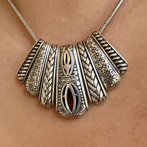 Puede incluir: Collar plateado con un colgante en forma de abanico. El colgante presenta intrincados dise&ntilde;os florales y trenzados, con un elemento central calado. El collar tiene una cadena delicada y est&aacute; dise&ntilde;ado para ser usado alrededor del cuello.