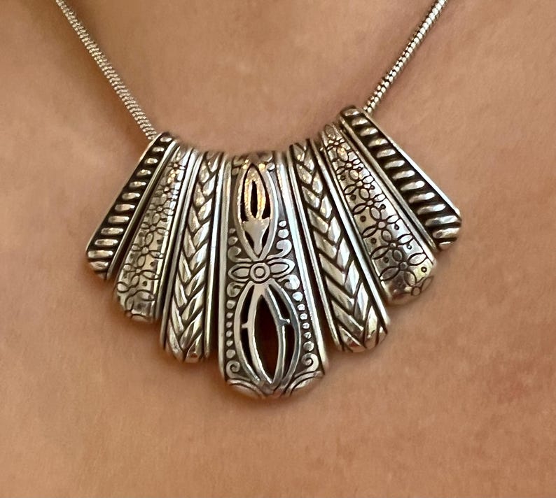 Puede incluir: Collar plateado con un colgante en forma de abanico. El colgante presenta intrincados dise&ntilde;os florales y trenzados, con un elemento central calado. El collar tiene una cadena delicada y est&aacute; dise&ntilde;ado para ser usado alrededor del cuello.