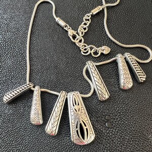 Puede incluir: Collar plateado con m&uacute;ltiples colgantes. Los colgantes presentan intrincados dise&ntilde;os florales y geom&eacute;tricos. El collar tiene una cadena de serpiente y una cadena ajustable con un dije en forma de coraz&oacute;n. La joya se exhibe sobre una superficie negra.