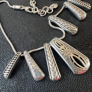 Puede incluir: Un collar plateado con m&uacute;ltiples colgantes. Los colgantes tienen varios patrones grabados, incluyendo dise&ntilde;os florales y geom&eacute;tricos. La cadena del collar es de estilo serpiente y el cierre es de mosquet&oacute;n.