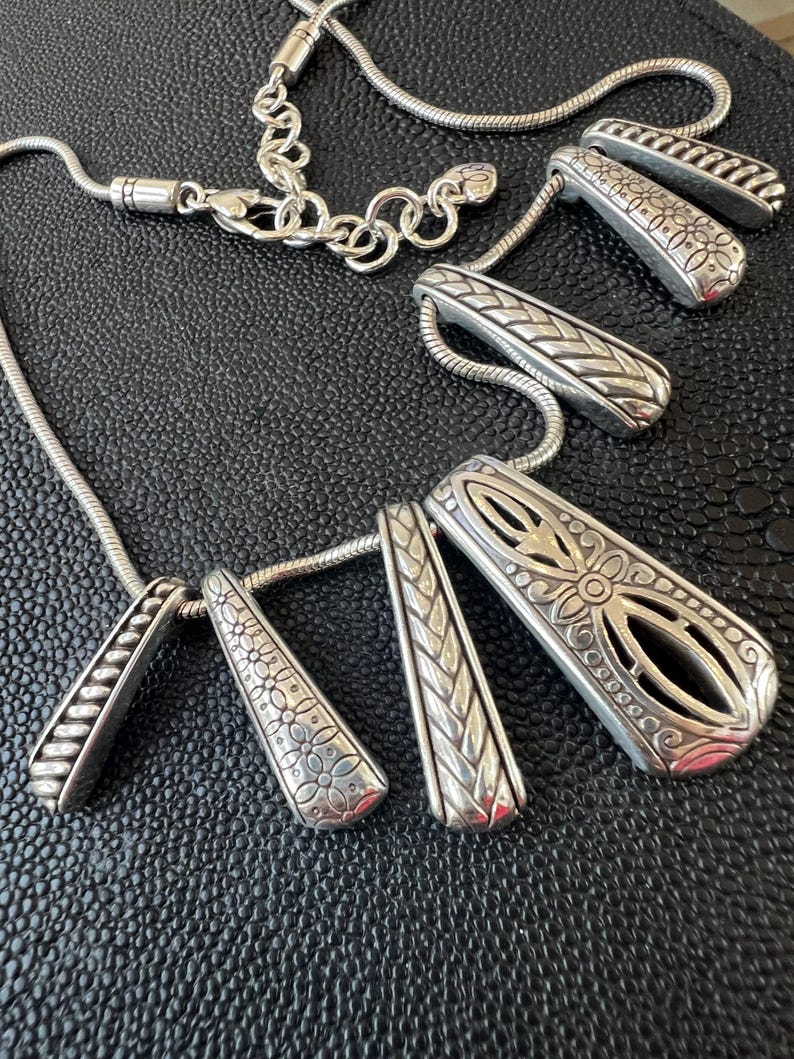 Puede incluir: Un collar plateado con m&uacute;ltiples colgantes. Los colgantes tienen varios patrones grabados, incluyendo dise&ntilde;os florales y geom&eacute;tricos. La cadena del collar es de estilo serpiente y el cierre es de mosquet&oacute;n.