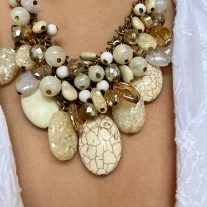 May include: A statement necklace featuring a mix of cream, beige, and brown beads and pendants. The necklace includes round, oval, and irregular-shaped beads, with a gold-tone chain.