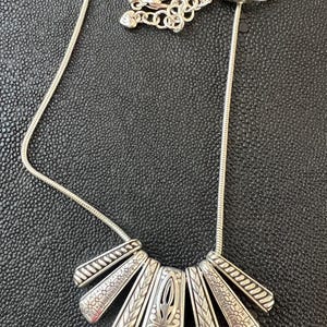 Puede incluir: Collar plateado con un colgante en forma de abanico. El colgante presenta un dise&ntilde;o central calado con motivos florales y geom&eacute;tricos. El collar tiene una cadena delicada y un cierre de mosquet&oacute;n. El colgante mide unos 5 cm de ancho.