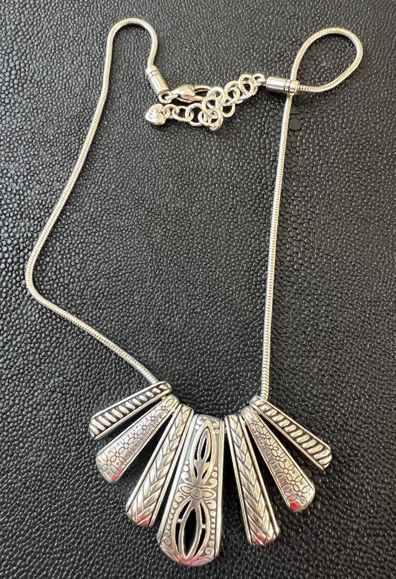 Puede incluir: Collar plateado con un colgante en forma de abanico. El colgante presenta un dise&ntilde;o central calado con motivos florales y geom&eacute;tricos. El collar tiene una cadena delicada y un cierre de mosquet&oacute;n. El colgante mide unos 5 cm de ancho.