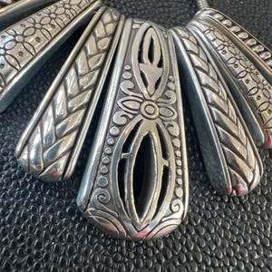 Puede incluir: Un collar plateado con un colgante en forma de abanico. El colgante presenta intrincados patrones florales y geom&eacute;tricos, con un dise&ntilde;o central abierto. El collar tiene una cadena de serpiente y se muestra sobre una superficie negra texturizada.