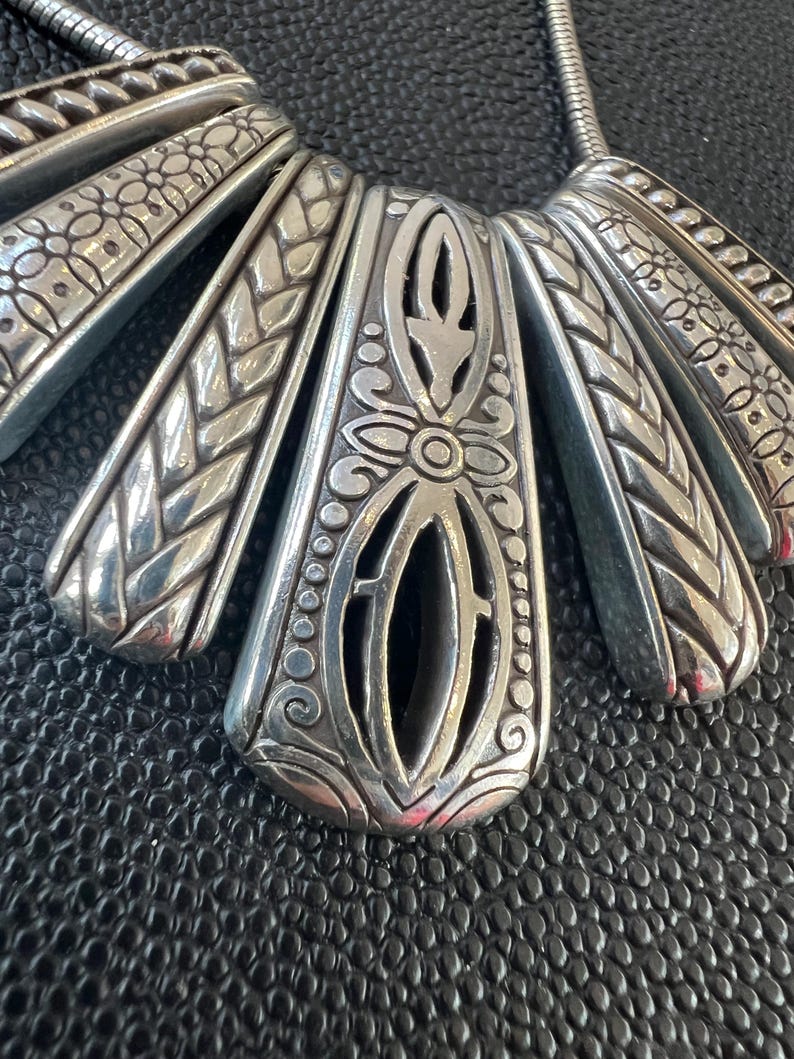 Puede incluir: Un collar plateado con un colgante en forma de abanico. El colgante presenta intrincados patrones florales y geom&eacute;tricos, con un dise&ntilde;o central abierto. El collar tiene una cadena de serpiente y se muestra sobre una superficie negra texturizada.