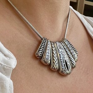 Puede incluir: Collar plateado con un colgante en forma de abanico. El colgante presenta intrincados patrones florales y geom&eacute;tricos, con un dise&ntilde;o calado central. El collar tiene una cadena delicada y se lleva alrededor del cuello.