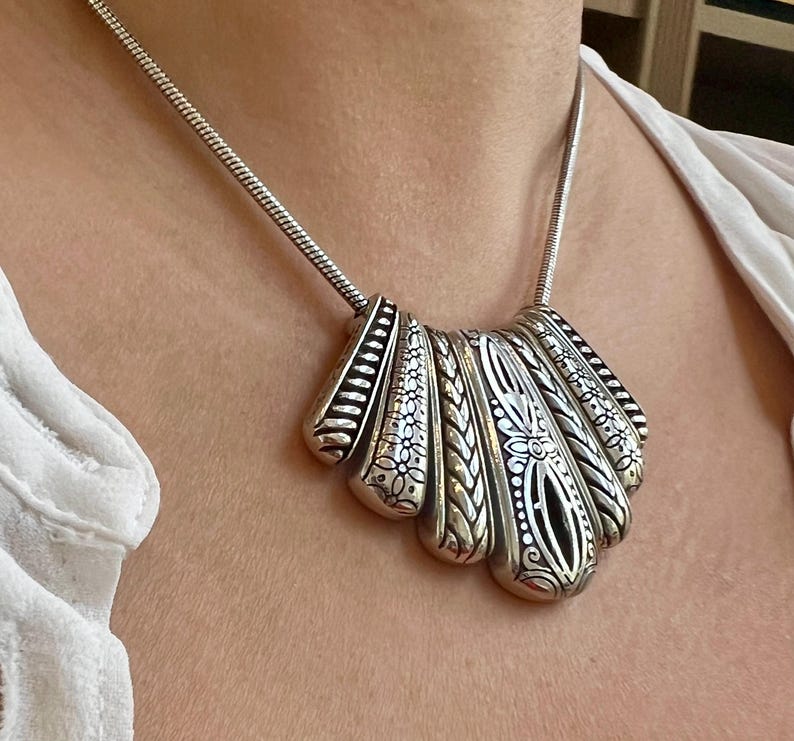 Puede incluir: Collar plateado con un colgante en forma de abanico. El colgante presenta intrincados patrones florales y geom&eacute;tricos, con un dise&ntilde;o calado central. El collar tiene una cadena delicada y se lleva alrededor del cuello.