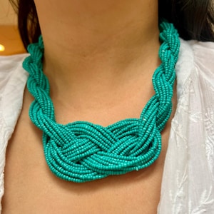 May include: A turquoise beaded necklace with a braided design. The necklace is made of small, round beads and is worn around the neck. The necklace is a vibrant shade of turquoise and is a statement piece.