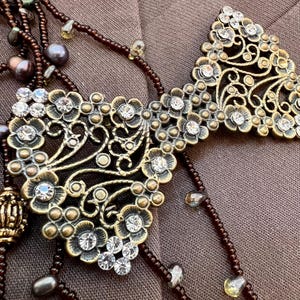 May include: An elaborate, vintage-style necklace with a bronze filigree pendant, embellished with clear crystals and small gold accents. The necklace includes dark brown beaded strands and pearls, creating a classic, antique look.