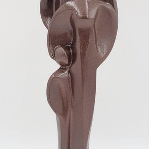 May include: A bronze-colored sculpture depicting two figures embracing. The abstract design features rounded shapes and a textured surface. The sculpture is mounted on a square base and stands against a white background.