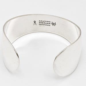 May include: A silver cuff bracelet with a smooth, polished finish. The interior is stamped with the text FAUSING STERLING DENMARK, a symbol resembling the letter A with a circle above it, and a stylized monogram logo featuring the letters W and A.