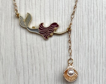 May include: Gold necklace with a mermaid charm and a pearl pendant. The mermaid charm is red, gray, and white. The pearl pendant is in a seashell.