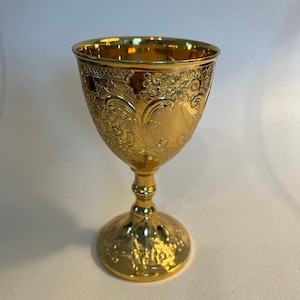 May include: A gold-colored chalice with an ornate design. The chalice has a round base and a tall stem.