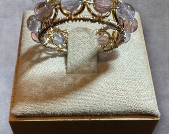 Puede incluir: Anillo abierto hecho a mano con un diseño de alambre dorado. El anillo está adornado con una serie de cuentas translúcidas, de color púrpura claro y transparentes. El anillo se exhibe en un expositor de joyas de madera con superficie de tela beige.