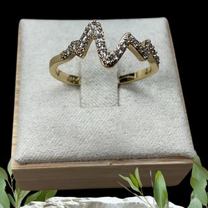 May include: A gold ring featuring a zigzag pattern, adorned with small, clear gemstones. The ring is presented on a light-coloured, textured surface inside a wooden box, set against a black backdrop. The ring's design resembles a heartbeat.