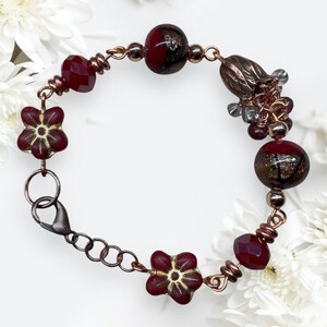 May include: A copper bracelet with burgundy faceted beads, flower-shaped beads, and round beads with gold flecks. The bracelet has a lobster clasp and a cluster of small beads. The bracelet is approximately 18 cm in circumference.