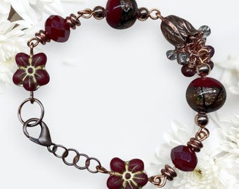 Pode incluir: Uma pulseira cor de cobre com contas facetadas bordô, contas em forma de flor e contas redondas com manchas douradas. A pulseira tem um fecho lagosta e um grupo de pequenas contas.