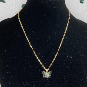 May include: A gold-coloured chain necklace featuring a butterfly pendant. The pendant has a translucent, iridescent appearance, with green and blue hues. The necklace is displayed on a black mannequin.
