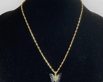 Pode incluir: Um colar de corrente dourada com um pingente de borboleta. O pingente de borboleta tem uma aparência translúcida e iridescente, com tons de verde e azul. O colar é exibido em um manequim preto.