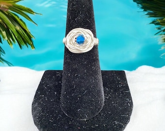 Puede incluir: Un anillo de alambre plateado con un diseño tejido, centrado con una gema azul vibrante. El anillo se exhibe en un soporte negro para anillos, con un fondo de una escena tropical.