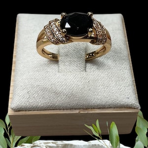 May include: A gold ring with a large, oval black gemstone. The band is embellished with small, clear gemstones. The ring is presented on a light-coloured, textured surface.