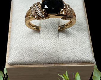 Pode incluir: Um anel dourado com uma grande pedra preta oval. A banda é adornada com pequenas pedras preciosas transparentes. O anel é apresentado numa superfície clara e texturizada.