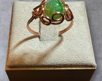 Puede incluir: Un anillo de tono cobrizo con una vibrante piedra verde en forma de esfera. El diseño del anillo incluye intrincados trabajos de alambre alrededor de la piedra y una banda sencilla. El anillo se exhibe en una caja de exhibición de joyas beige cubierta de tela.