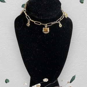 May include: A gold-coloured chain bracelet with a rectangular link design. The bracelet features a cat-shaped charm and several dangling charms, including small round gems. The bracelet is displayed on a black jewellery bust.