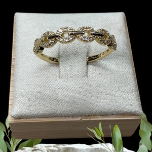 May include: A gold ring featuring a chain link design, adorned with small, clear stones. The ring is presented on a light-coloured fabric within a wooden box, set against a black backdrop.